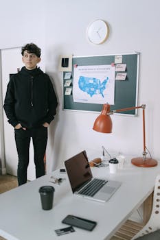 Young professional standing in a modern office with tech devices and a map on the wall.